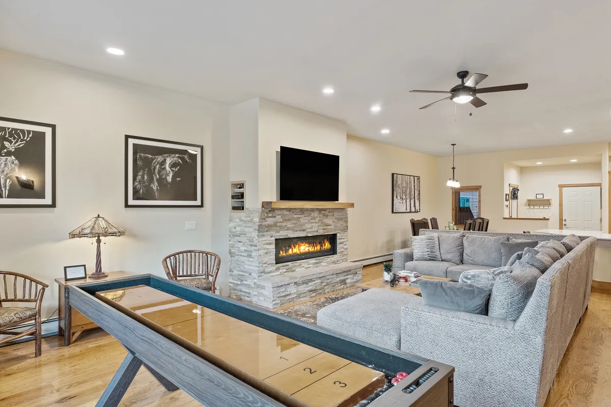 An inviting living space showing a cozy fireplace, comfortable sofa, and prominently featuring a shuffleboard table set up for play. Artwork and soft lighting add warmth, while the arrangement encourages both relaxation and playful interaction, embodying the spirit of family game nights and group fun.