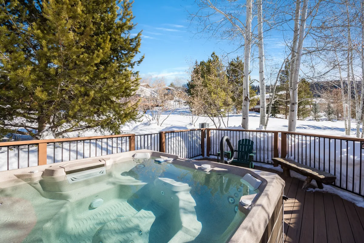 A steaming community hot tub sits on a deck surrounded by snow, framed by trees and a snow-blanketed landscape. The setting is tranquil and inviting, offering the quintessential après-ski experience for guests looking to unwind after skiing or snowboarding.