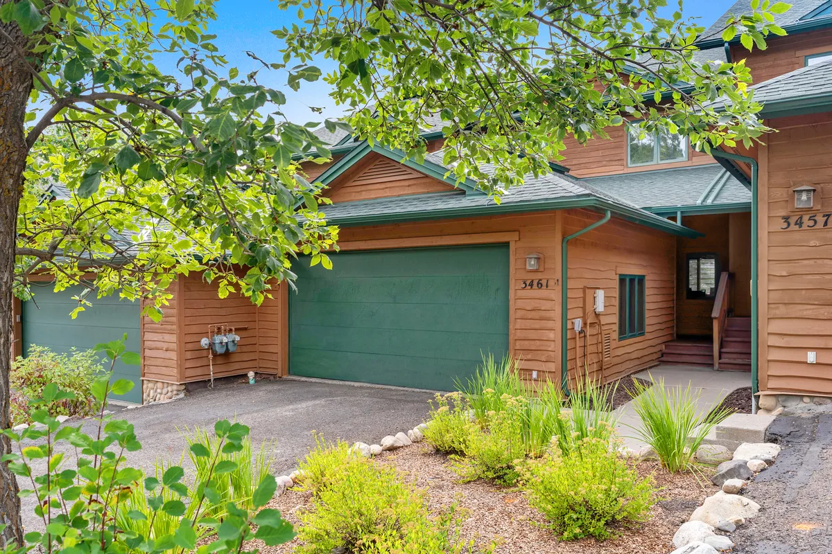 This image features the exterior of the home with a green garage door, wooden siding, lush greenery, and a stone border. The welcoming front deck is visible, bathed in sunlight and surrounded by summer foliage, creating an inviting entrance ideal for enjoying a morning coffee or gathering with friends. The oversized garage is showcased as convenient for storing adventure gear.