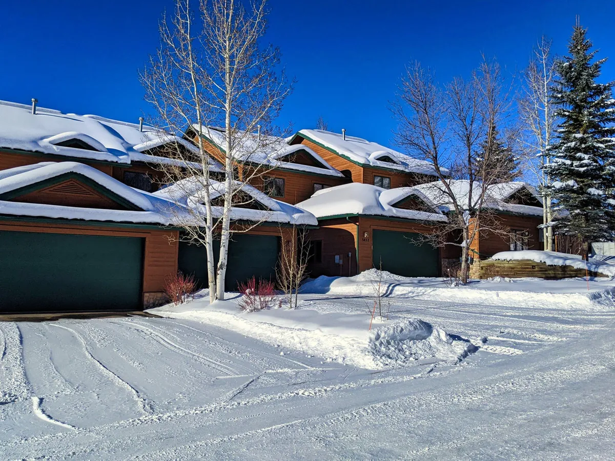 Another exterior perspective of the snow-covered townhome, showcasing the wooden siding, green garage doors, and bare winter trees. The image highlights both the home’s proximity to nature and its welcoming ambiance amid the winter landscape.