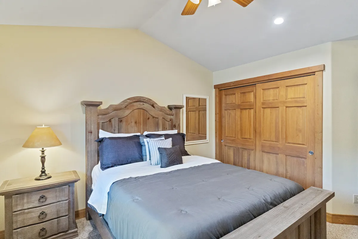 This inviting bedroom features a wooden headboard, cozy bedding, a nightstand with a lamp, and closet doors. Soft light and decorative pillows enhance the feeling of warmth and relaxation.