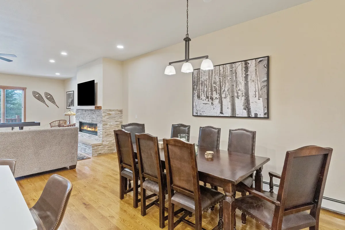 A warm, inviting dining area with a large table set for eight, complemented by a modern light fixture, a striking fireplace, and nature-inspired artwork. The space exudes a sense of celebration and togetherness, ideal for group meals or festive occasions.