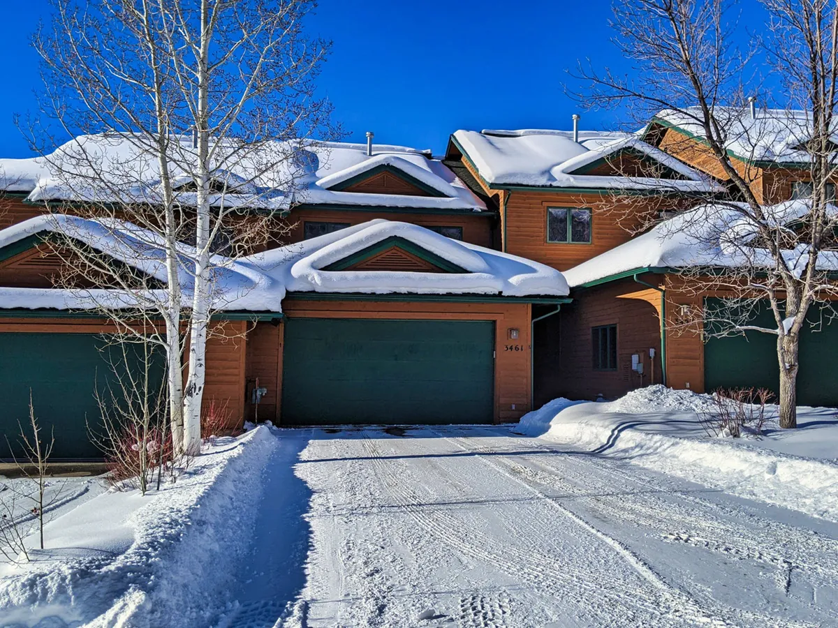 A picturesque exterior view of the townhome with snow-covered roofs and green garage doors, surrounded by tall trees dusted with snow. The scene evokes a classic winter atmosphere, perfect for families or groups returning from a day on the slopes. The warm glow from inside contrasts beautifully with the crisp outdoor setting, hinting at cozy moments after outdoor adventures.