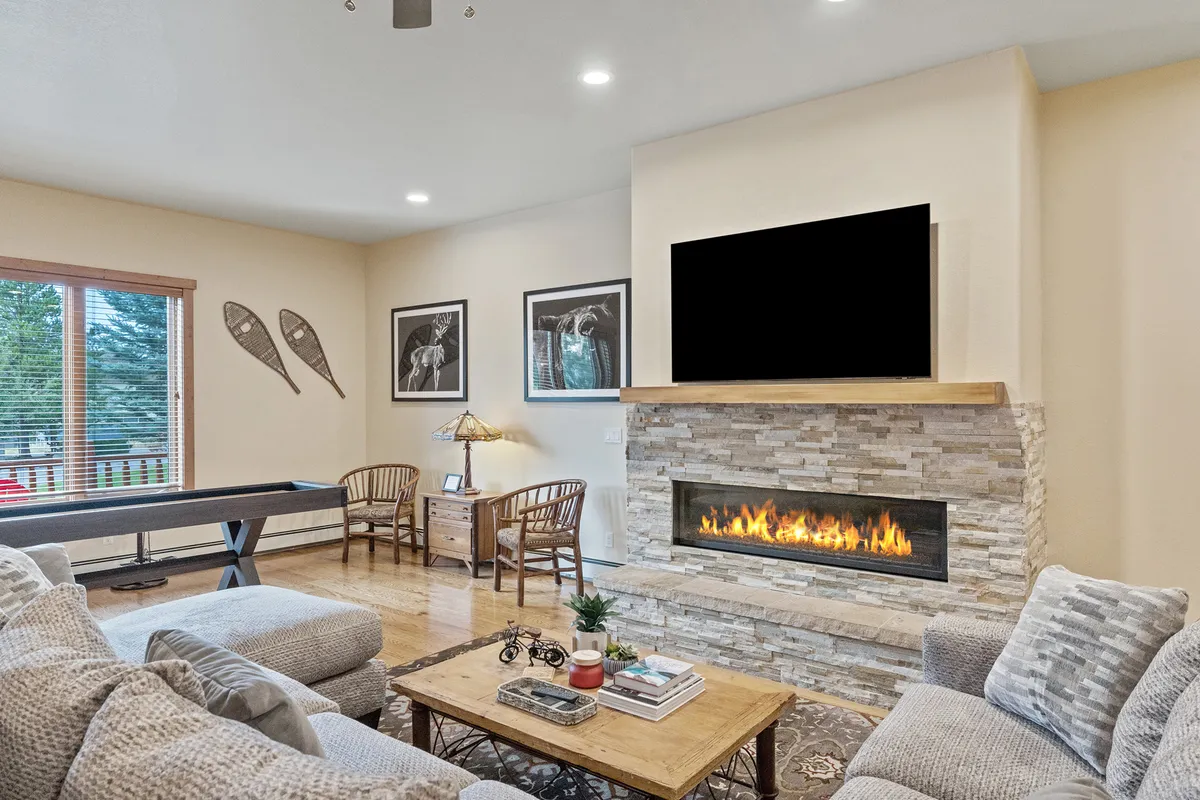 A cozy, open-concept living area featuring a modern fireplace, plush sofa, eye-catching artwork, and a spacious dining table next to large windows that let in plenty of natural light. The space invites groups to gather for games, conversations, or relaxing evenings.