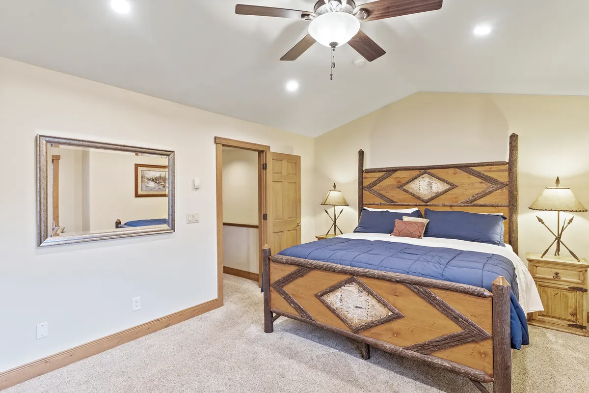 A tranquil main bedroom with a neatly made bed, decorative mirror, matching nightstands, and warm, inviting lamps. Natural light pours in from a window, illuminating the workspace area and highlighting the calming ambiance.