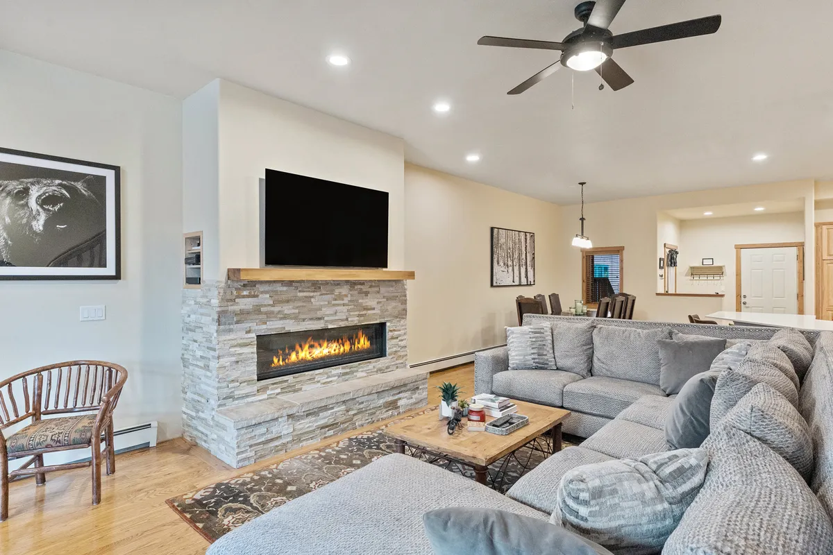 A warm, open living room featuring a large comfortable sectional sofa centered around a sleek modern gas fireplace. The space is styled with inviting decor, a wooden coffee table, and artwork on the walls, evoking a sense of togetherness and relaxation. The overall mood is welcoming, perfect for gathering with family or friends.